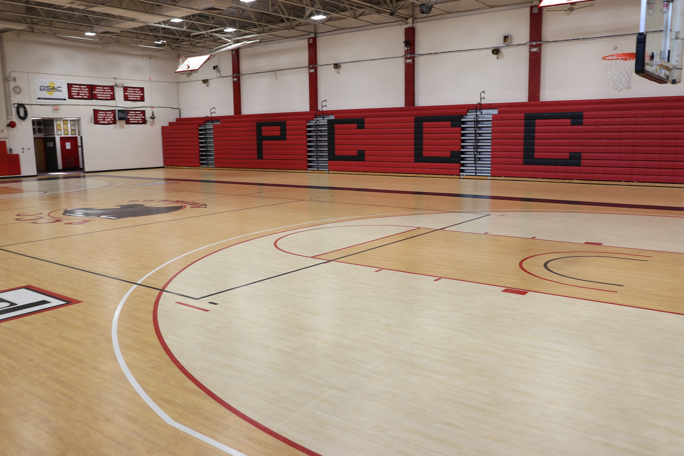 Empty indoor basketball gymnasium with a polished wood court, two basketball hoops, and red retractable bleachers along one wall. Large black letters are displayed across the bleachers, and school banners hang on the walls above the seating area. The gym is brightly lit by overhead lights and has no people present.