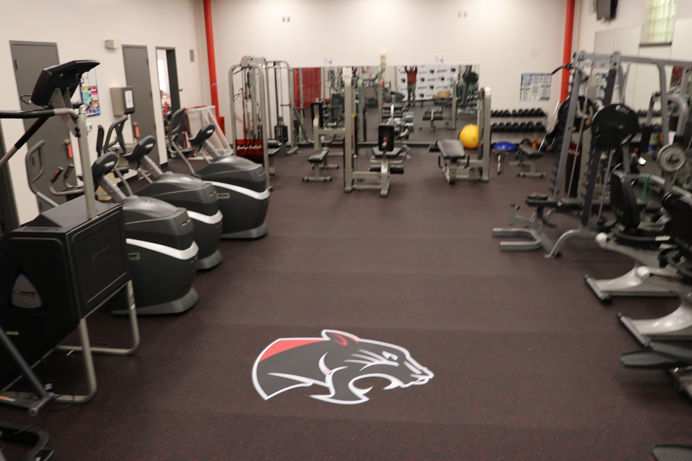 Spacious athletic fitness center equipped with cardio machines, weight-training equipment, free weights, and mirrored walls. The facility features rubberized flooring with a panther logo at center, providing a dedicated space for strength training and athletic conditioning.