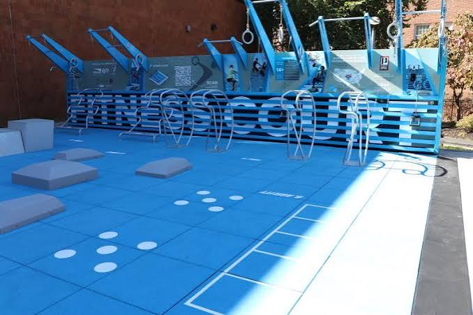Outdoor fitness and conditioning area with bright blue training surfaces, obstacle-course elements, climbing structures, gymnastic rings, balance platforms, and agility markings. The space is designed for functional fitness, strength training, and athletic conditioning activities.
